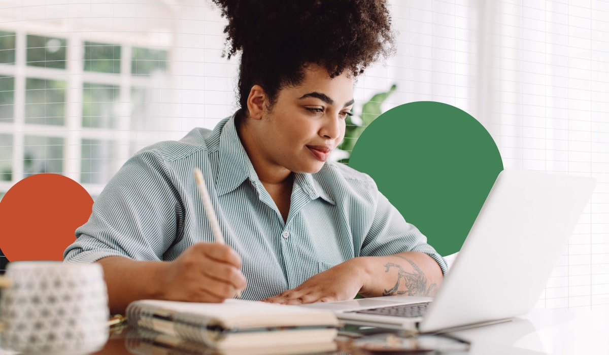 A woman sat at a desk, writing in a notebook while looking at a laptop screen.