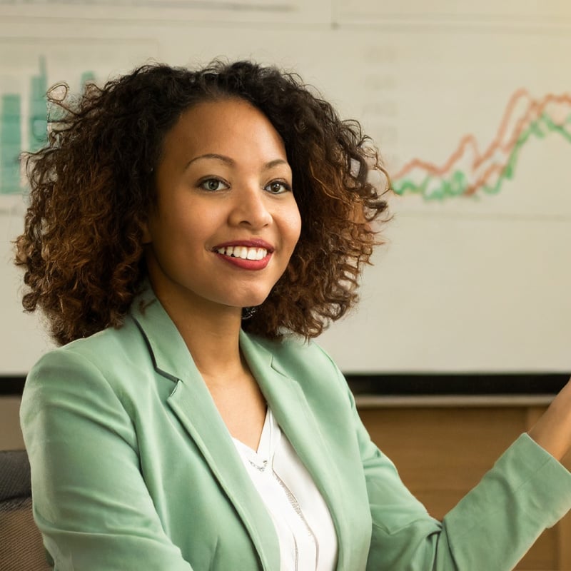 A Black woman smiling, wearing a light green blazer, standing in front of a series of financial charts and delivering a presentation