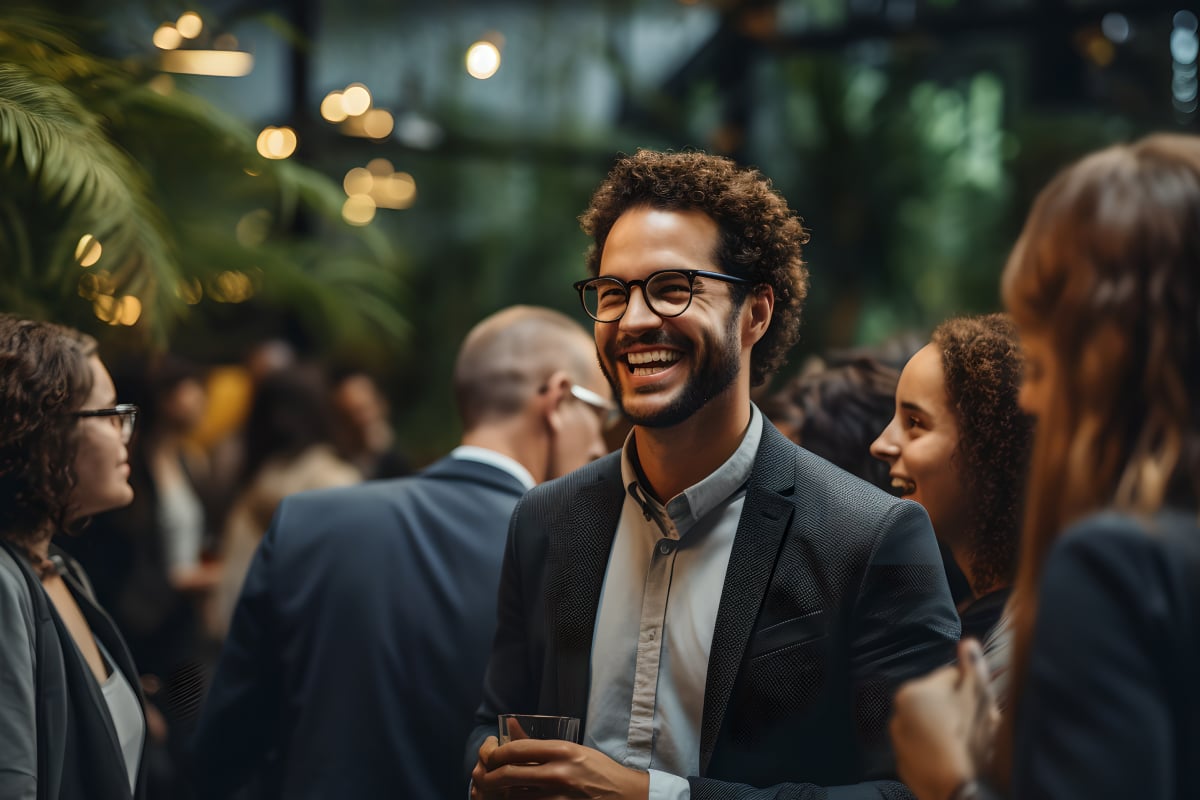 A man with olive skin, brown curly hair and a beard wearing glasses and professional attire smiling as he networks with other professionals at a conference