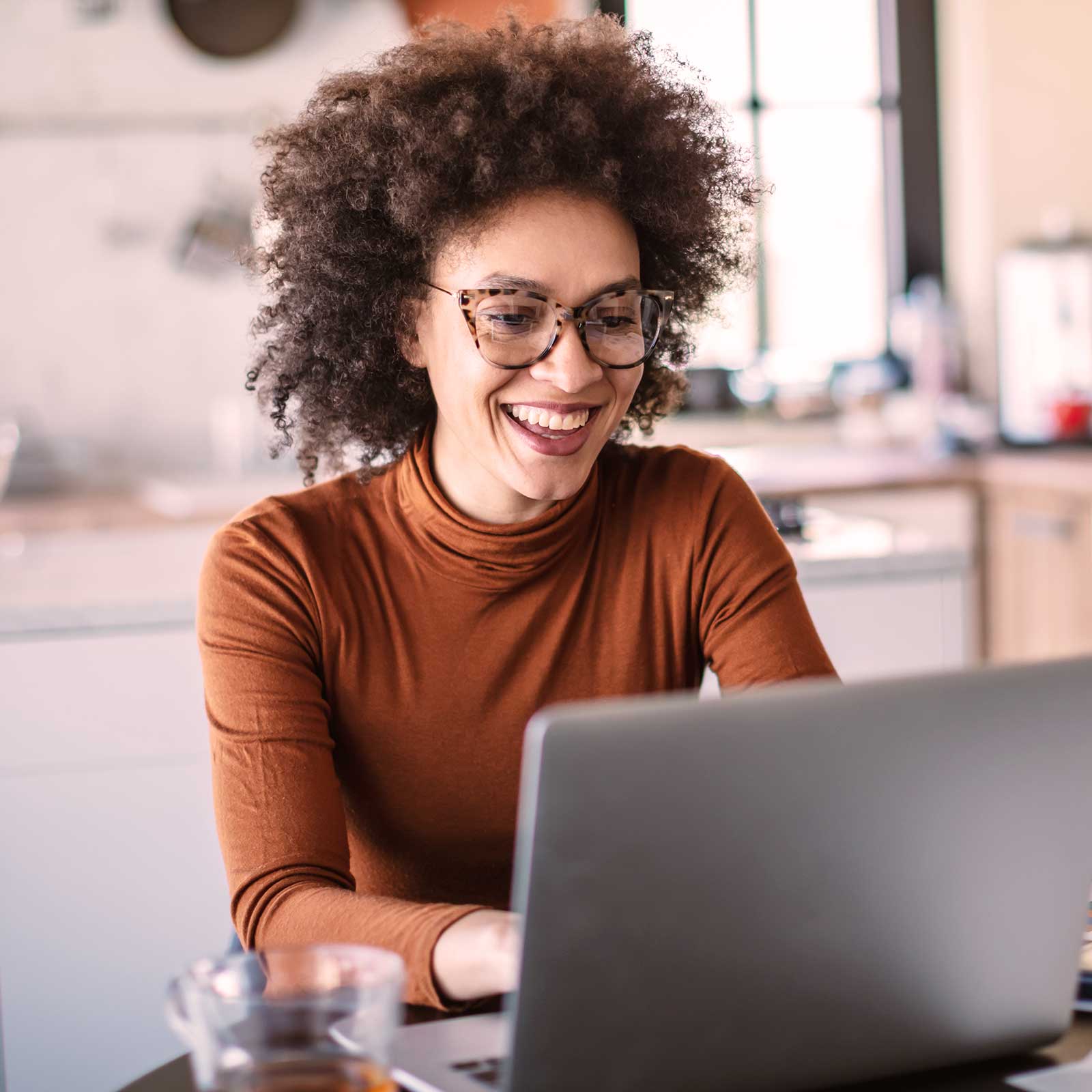 A smiling black woman with curly hair wearing a brown turtleneck sitting in front of a laptop working in the comfort of her home