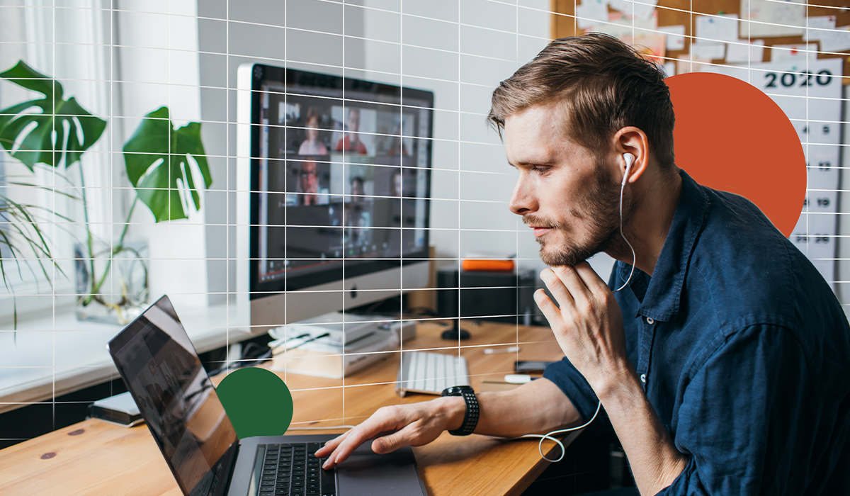 Man with earphones sitting at desk and looking at laptop with monitor in background.