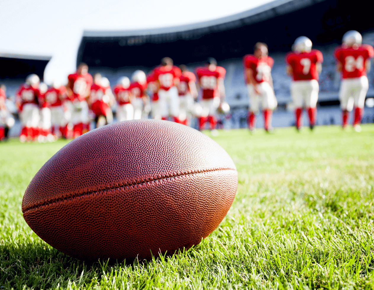 Photo of a football on a field with a team of players wearing red jerseys standing behind it in the background.