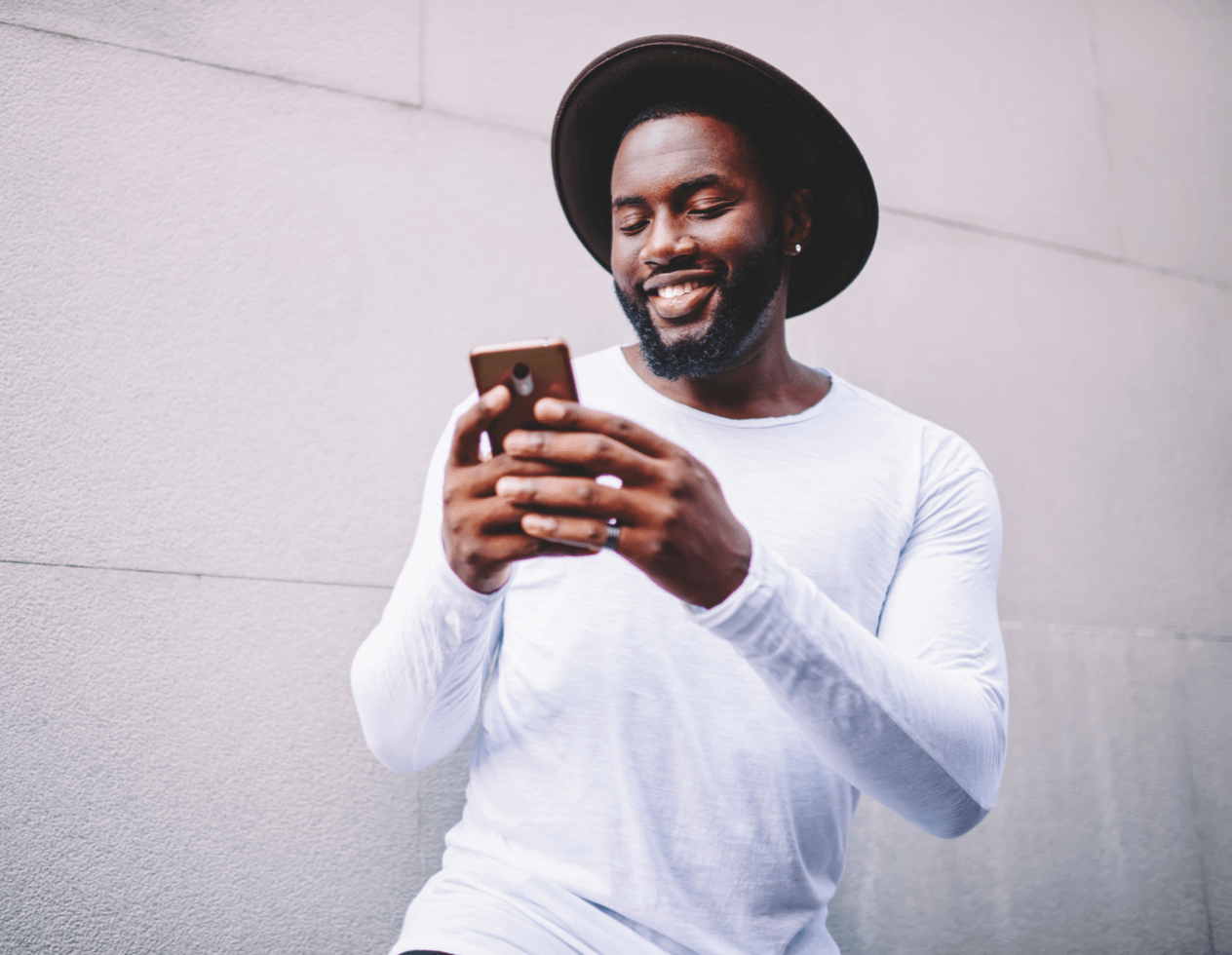 African American man wearing a white shirt and brown fedora smiling while looking at his cell phone.