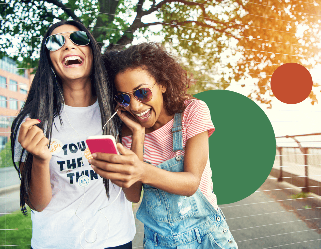 Two women in sunglasses laugh while walking down a street sharing headphones looking at a cell phone.