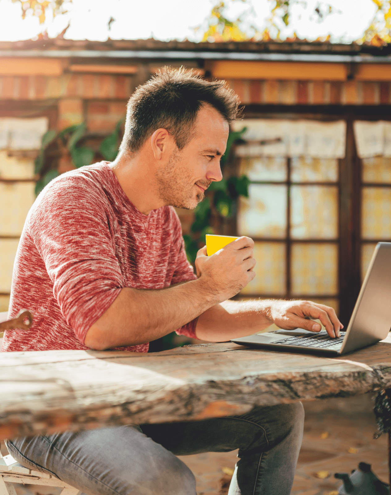 Caucasian man in a pink shirt working on a laptop computer while drinking a cup of coffee in his backyard.