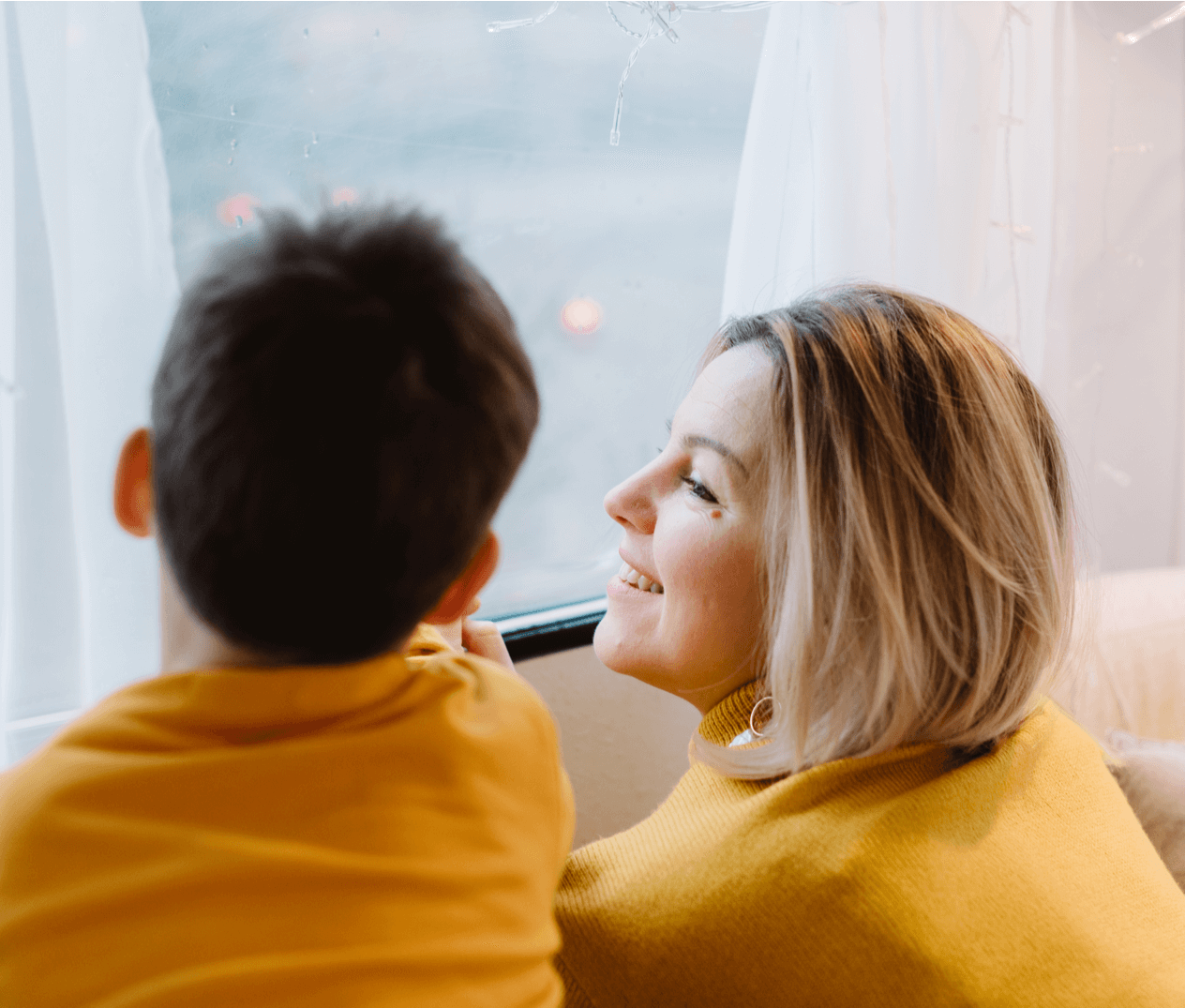 Caucasian woman smiling while looking out a window with her young son.