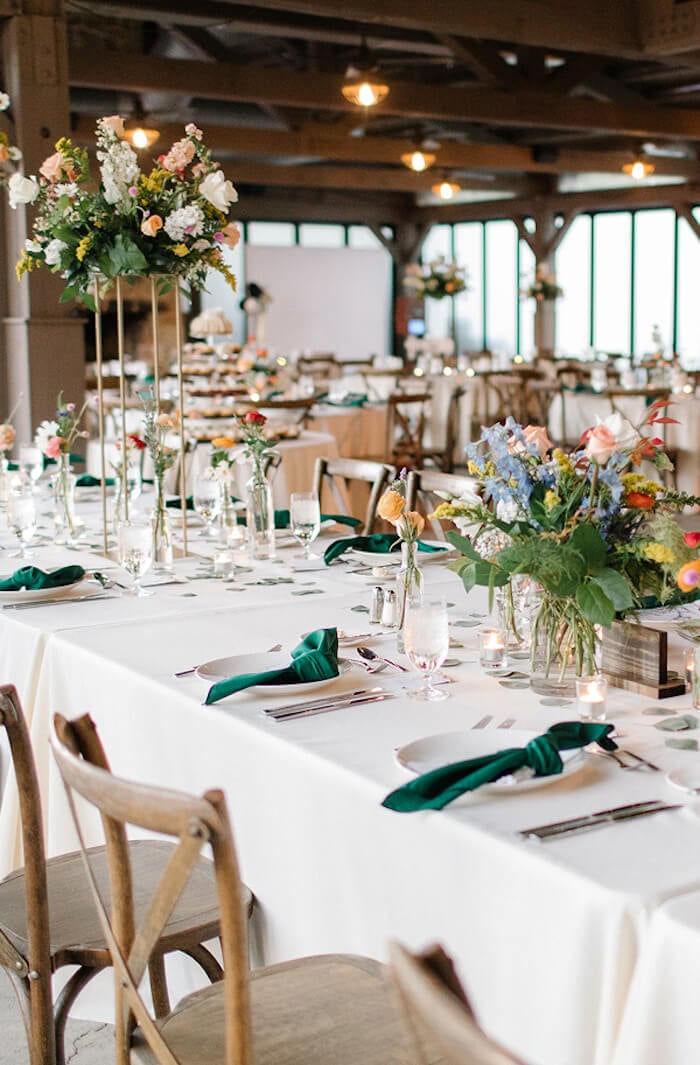 Long banquet tables with white linens, green napkins, wooden chairs, and tall floral centerpieces in a rustic venue