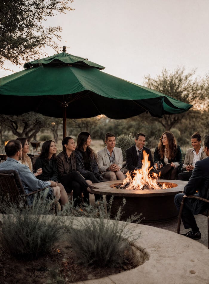 Group of people seated around an outdoor fire pit under a large green umbrella at dusk, chatting and smiling.