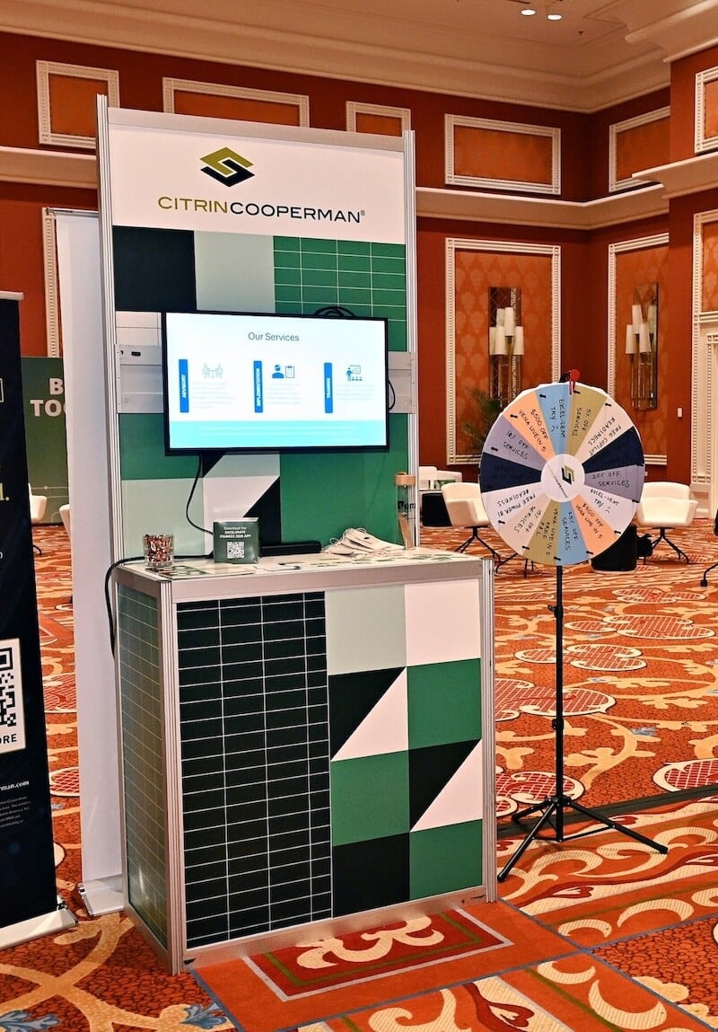 Citrin Cooperman trade show booth with “Our Services” screen and a prize wheel on a stand in a carpeted conference hall.