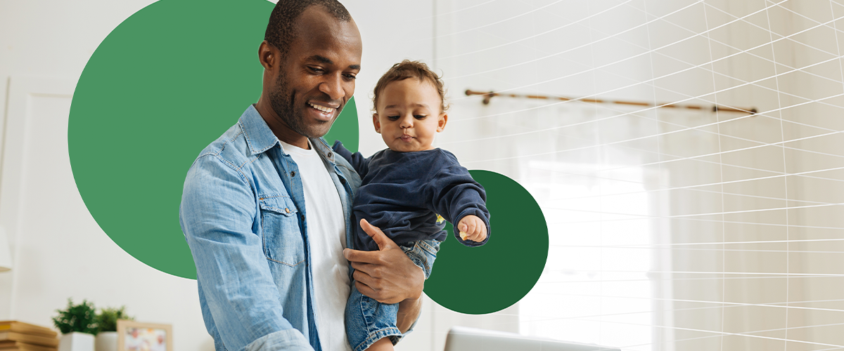 Smiling man holding a baby in one arm while typing on a laptop with his other hand.